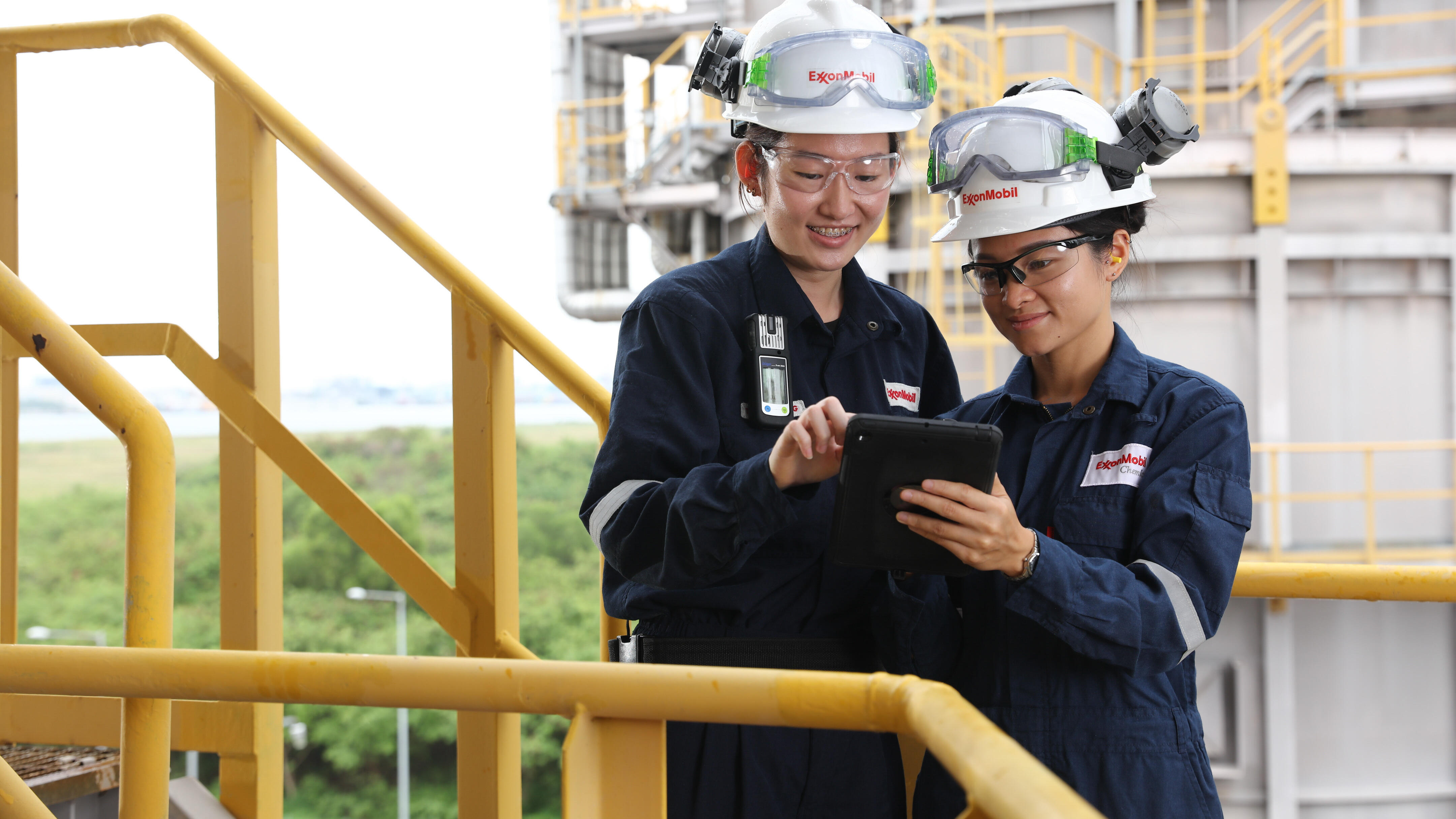 Two workers wearing navy blue coveralls and white safety helmets with the ExxonMobil logo, standing on a yellow metal staircase in an industrial setting. Both workers are equipped with safety goggles and appear to be engaged in a discussion while looking at a tablet device. The background includes industrial equipment and structures, with greenery visible beyond the facility. The environment suggests they are on a site related to energy, chemicals, or manufacturing, and they are likely reviewing operational or safety procedures.