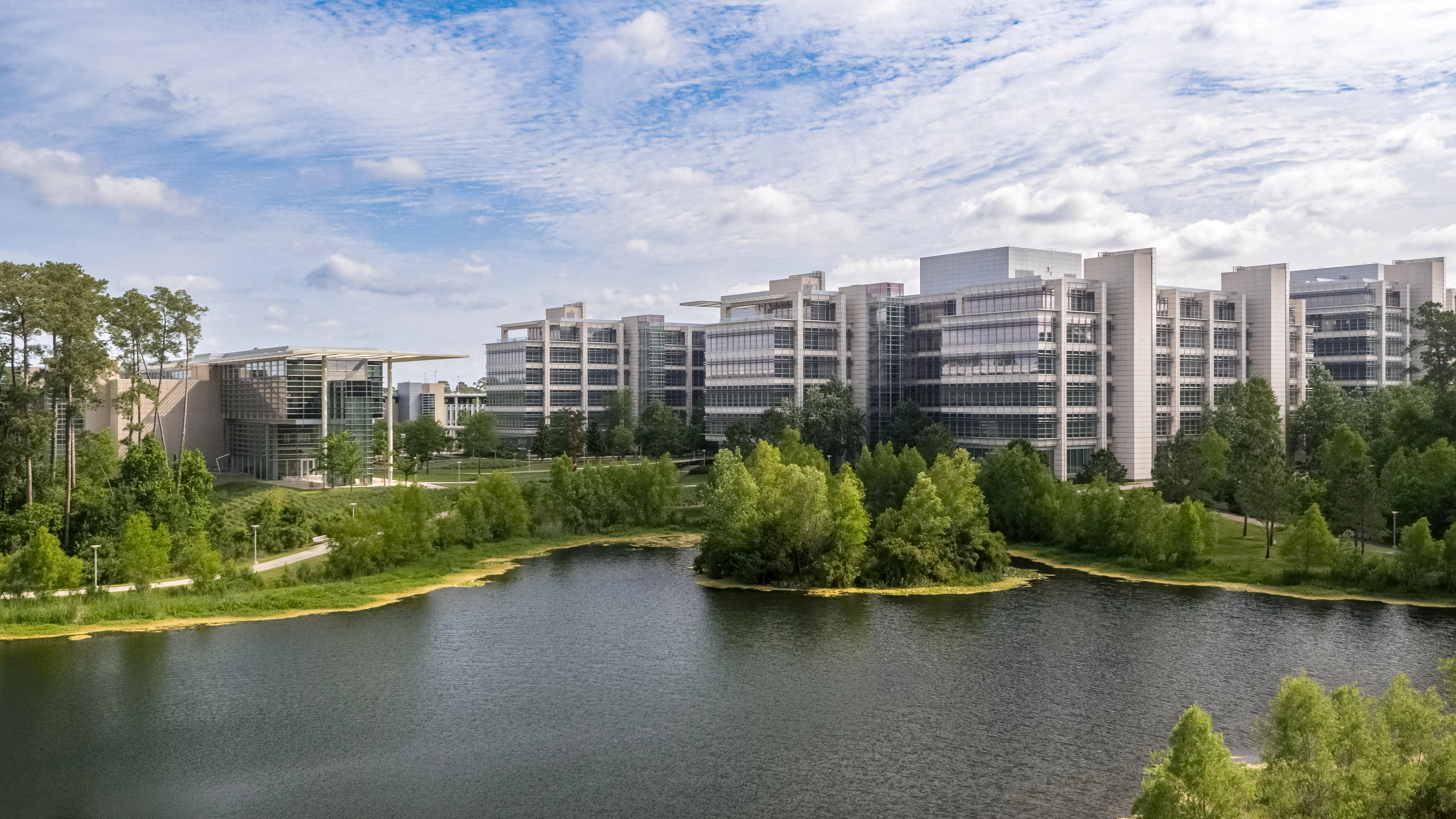 A modern office complex with several large, glass-fronted buildings situated behind a scenic pond. The architecture features clean lines and extensive use of windows, allowing natural light to flood the interiors. The foreground is dominated by the pond, surrounded by lush greenery and trees, providing a tranquil, park-like setting. The sky is partly cloudy, adding to the serene atmosphere. There are walking paths and outdoor spaces visible, suggesting the area is designed to encourage outdoor activity and relaxation.