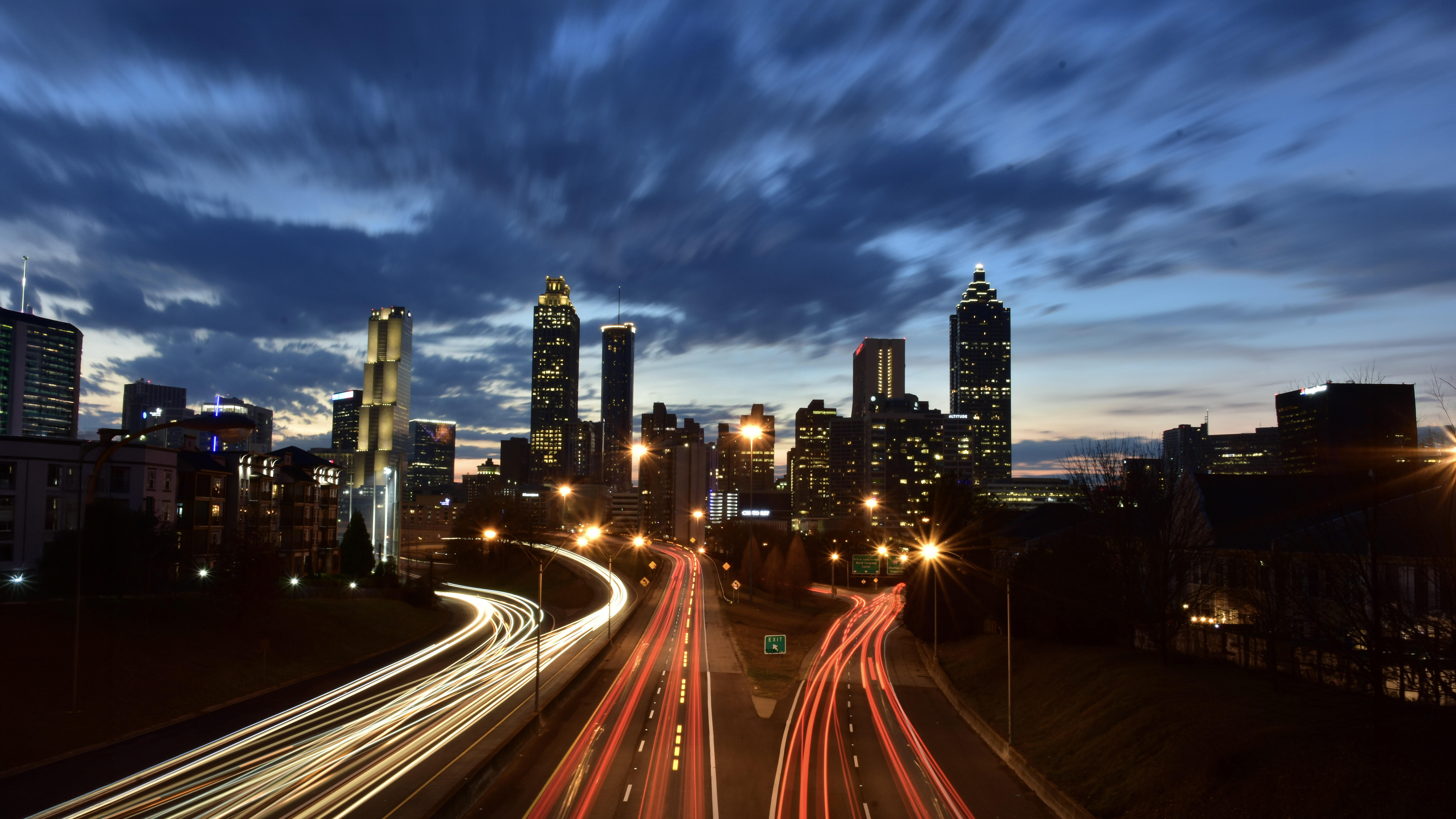 A dusk cityscape shows a modern skyline with tall, illuminated buildings under streaky blue clouds. In the foreground, a multi-lane highway curves into the city, captured with long exposure so headlights form white streams and taillights form red ribbons. Streetlights glow amber along the road, and the horizon still holds a faint band of sunset light. The scene feels dynamic and energetic as traffic flows toward the downtown core.