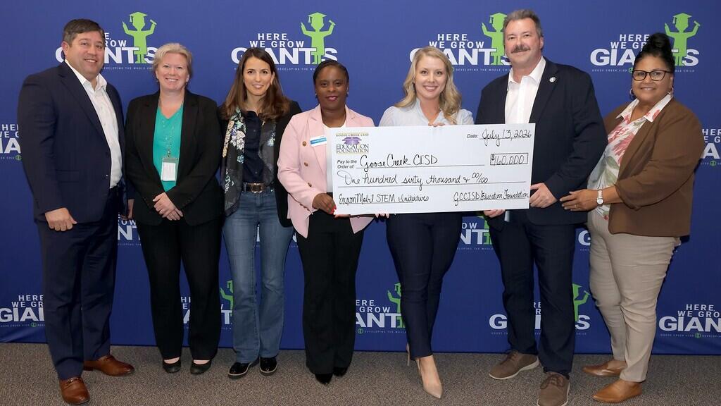 A group of eight adults poses in front of a blue backdrop that repeats the slogan “Here We Grow Giants.” Two people in the center hold an oversized check made out to a school district for $160,000, noting “Global STEM Education”